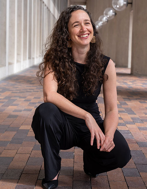 A color photograph of Guest Conductor Elizabeth Eschen Cacciola, who is smiling and kneeling, has white skin, long curly brown hair, and is wearing large gold earrings and black clothing.